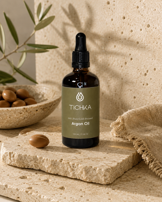 A bottle of Tichka Argan Oil serum in an amber glass dropper bottle with a muted olive-green label, styled on a textured stone slab with argan nuts and a linen cloth.