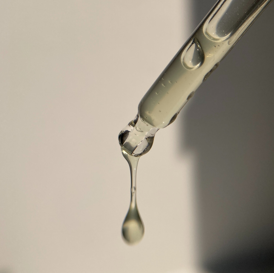 A close-up of a glass dropper tube with a single clear liquid drop hanging in the center against a soft gray background.