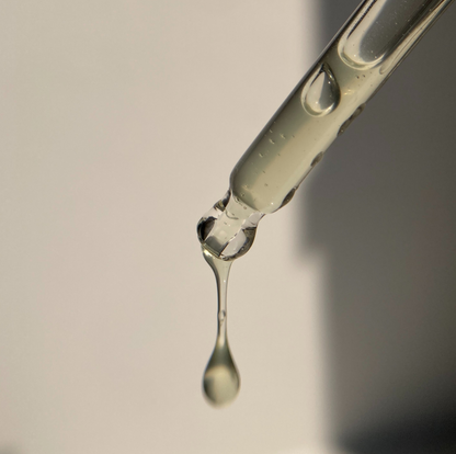 A close-up of a glass dropper tube with a single clear liquid drop hanging in the center against a soft gray background.