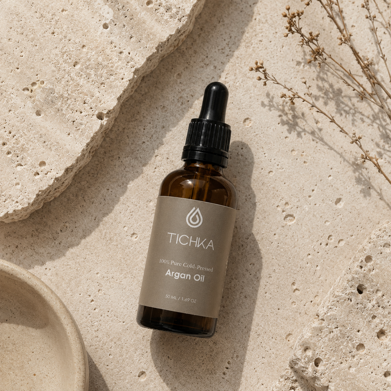 A bottle of TICHKA Argan Oil dropper bottle, placed on a textured beige stone surface with dried botanicals and a ceramic dish.