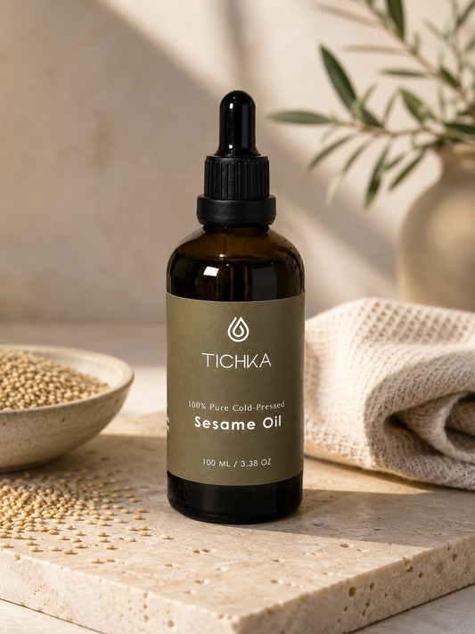 A 100ml amber glass dropper bottle of Tichka Pure Cold-Pressed Sesame Oil, styled on a stone slab with sesame seeds and a beige cloth.