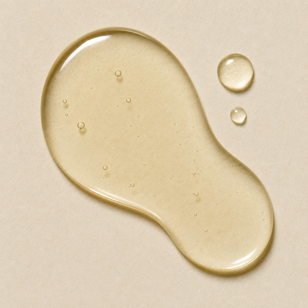 A glossy droplet of translucent pale yellow liquid on a warm beige background, with two smaller droplets nearby.