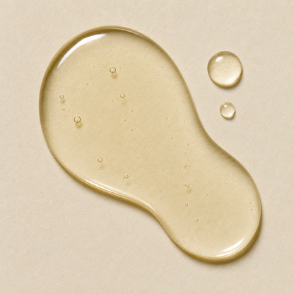 A glossy droplet of translucent pale yellow liquid on a warm beige background, with two smaller droplets nearby.