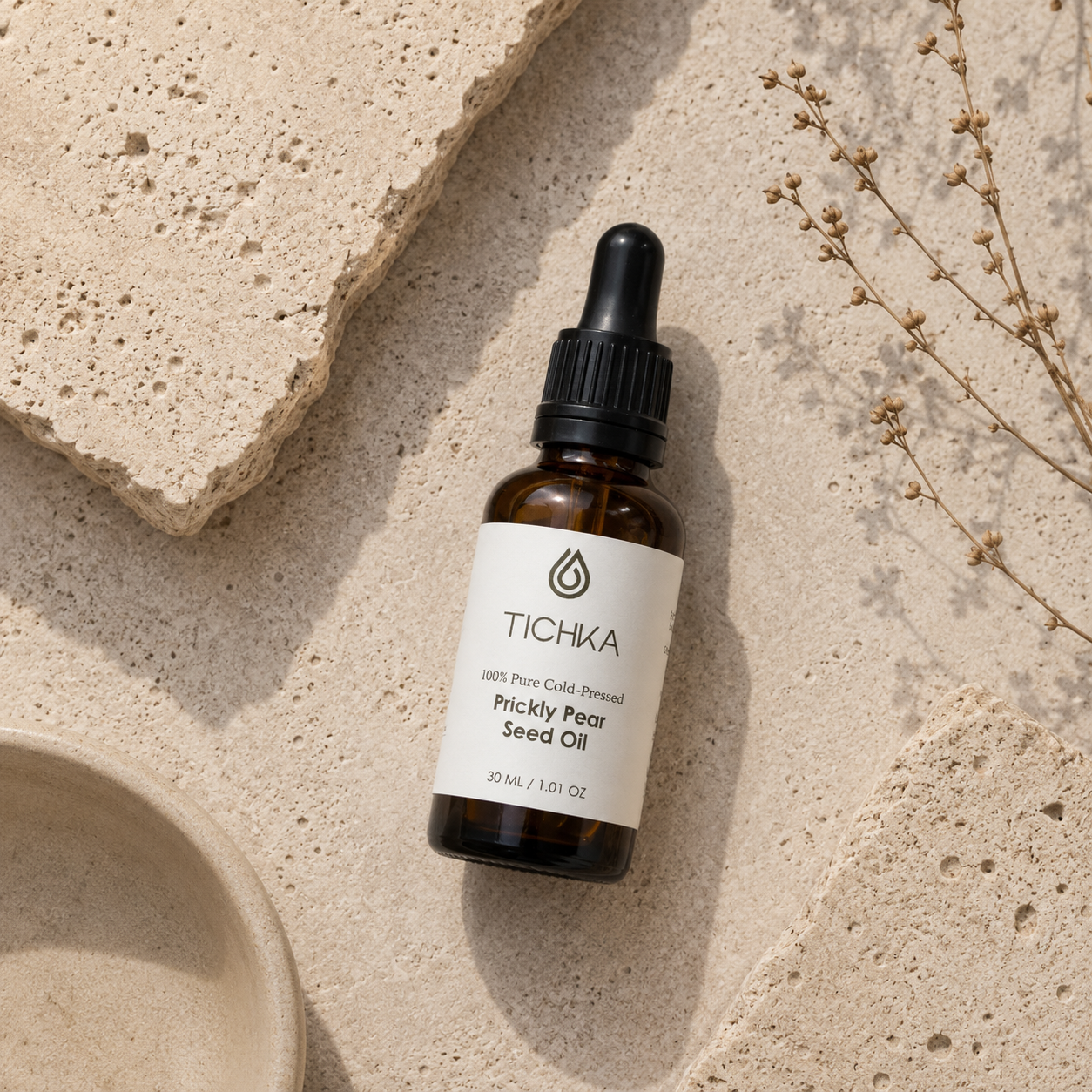 A small amber glass dropper bottle labeled TICHKA Prickly Pear Seed Oil, styled on a light stone surface with textured tiles and dried botanicals.