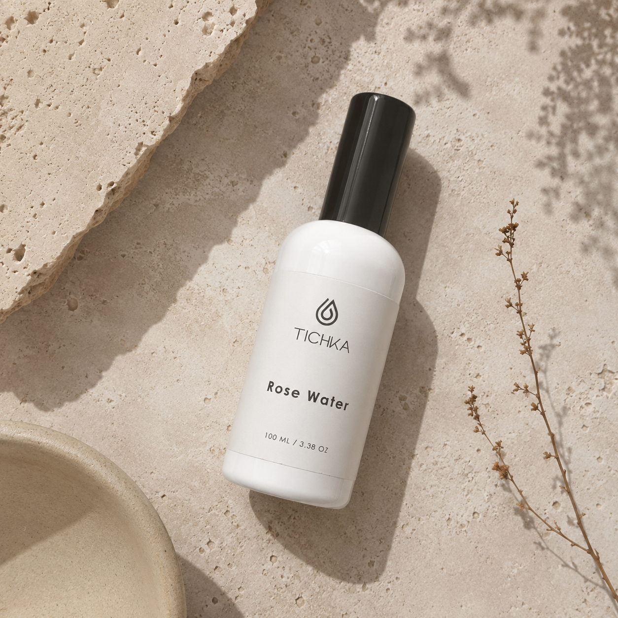 A white bottle of TICHKA Rose Water with a black cap, styled on a textured beige stone surface with a ceramic dish and dried plant sprigs.