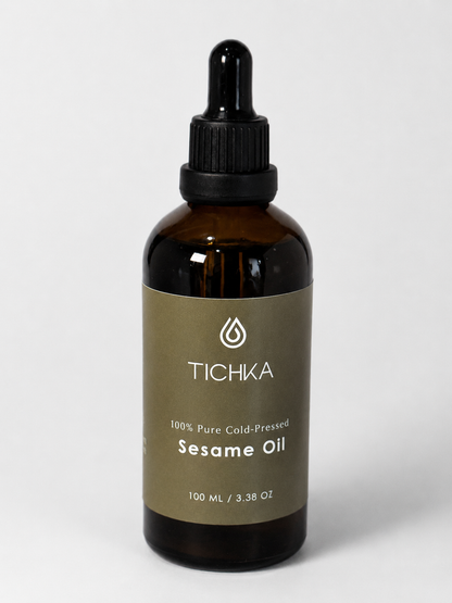 A 100 mL amber glass dropper bottle of Tichka Sesame Oil with a black dropper cap and a muted olive-brown label, set against a light gray background.